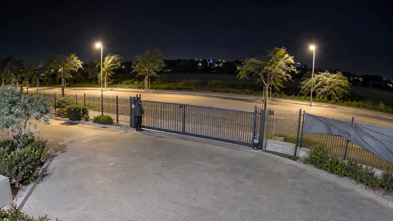 Entrée d'un site la nuit par grand vent : arbres, bâche et voiture bougent, puis une personne marche jusqu'au portail